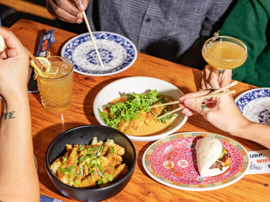 A group of diners sharing Asian-inspired dishes at a wooden table, with bao buns, French fries and roasted mushroom dumplings. Hands holding chopsticks reach to grab food from the plates of food.