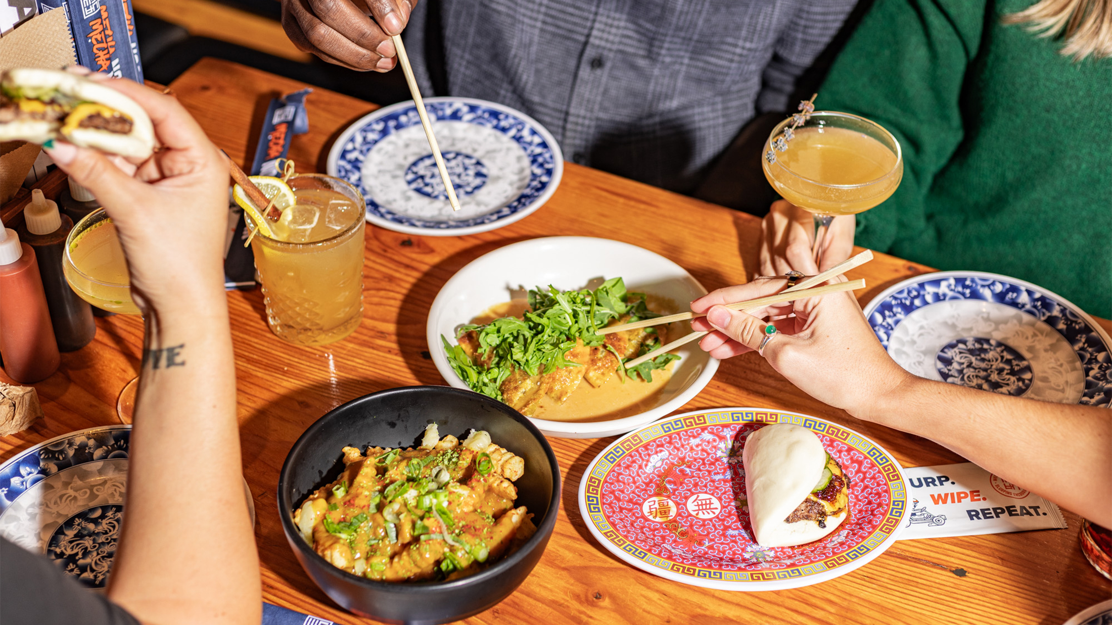 A group of diners sharing Asian-inspired dishes at a wooden table, with bao buns, French fries and roasted mushroom dumplings. Hands holding chopsticks reach to grab food from the plates of food.