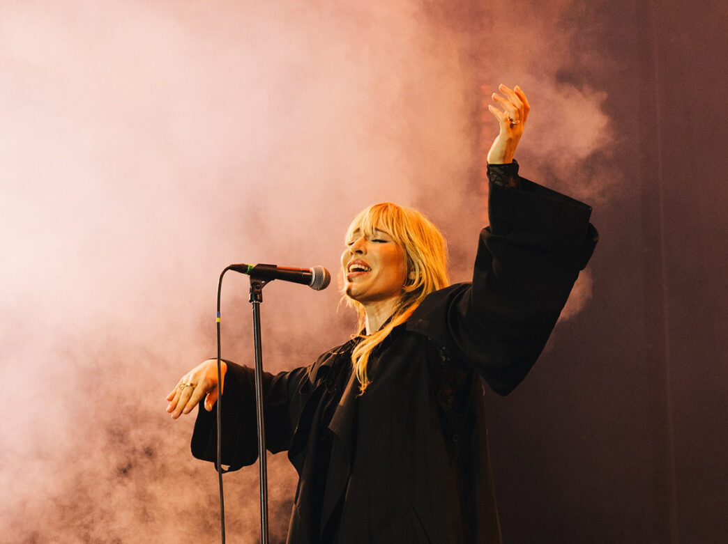 A blonde woman sings passionately into a microphone, with one arm raised in the air, as light pink stage smoke surrounds her.