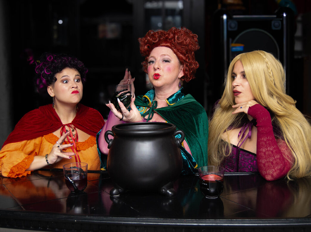 Three people dressed up as the witches from Hocus Pocus sit at a bar with a black cauldron, each posing dramatically in costume.