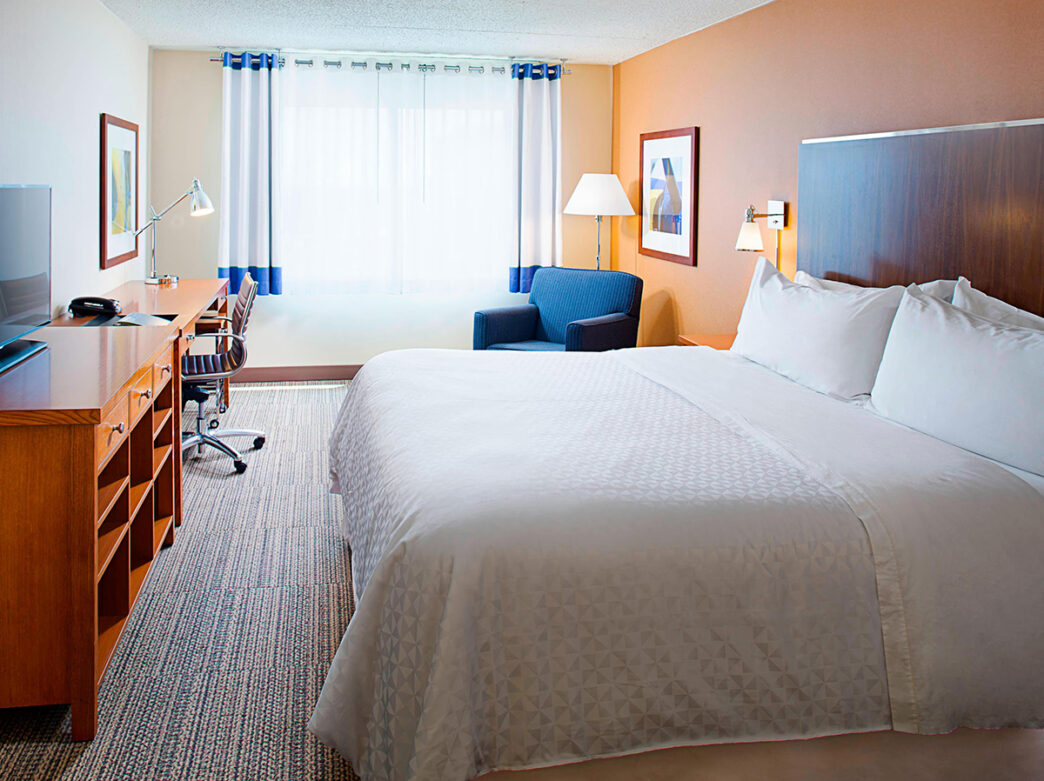 A bright hotels guestroom with a king bed, wooden desk, office chair and a lounge chair by the window.
