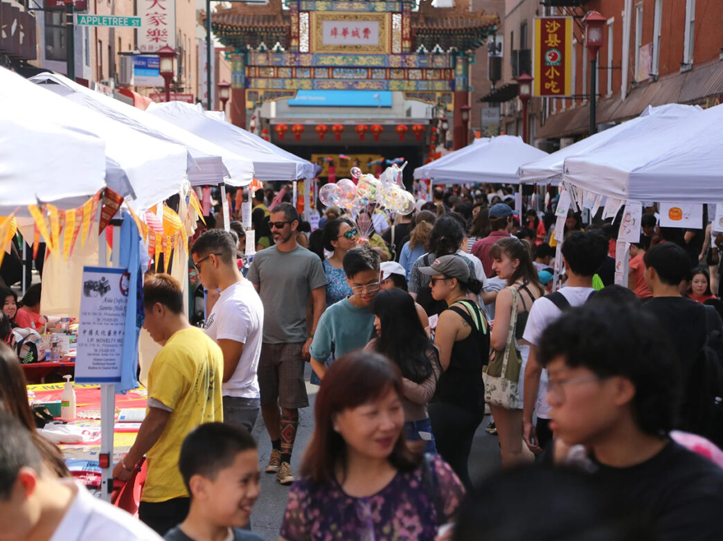 Crowds explore vendor tents along a busy, car-free street in Chinatown, with the colorful Friendship Gate in the background.