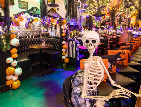 A plastic skeleton sits on a bar stool at a long wooden bar, surrounded by glowing pumpkin string lights, cobwebs, and colorful Halloween decorations hanging from the ceiling.