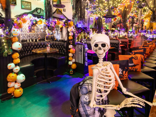A plastic skeleton sits on a bar stool at a long wooden bar, surrounded by glowing pumpkin string lights, cobwebs, and colorful Halloween decorations hanging from the ceiling.