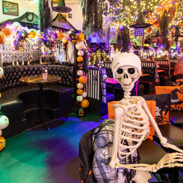 A plastic skeleton sits on a bar stool at a long wooden bar, surrounded by glowing pumpkin string lights, cobwebs, and colorful Halloween decorations hanging from the ceiling.