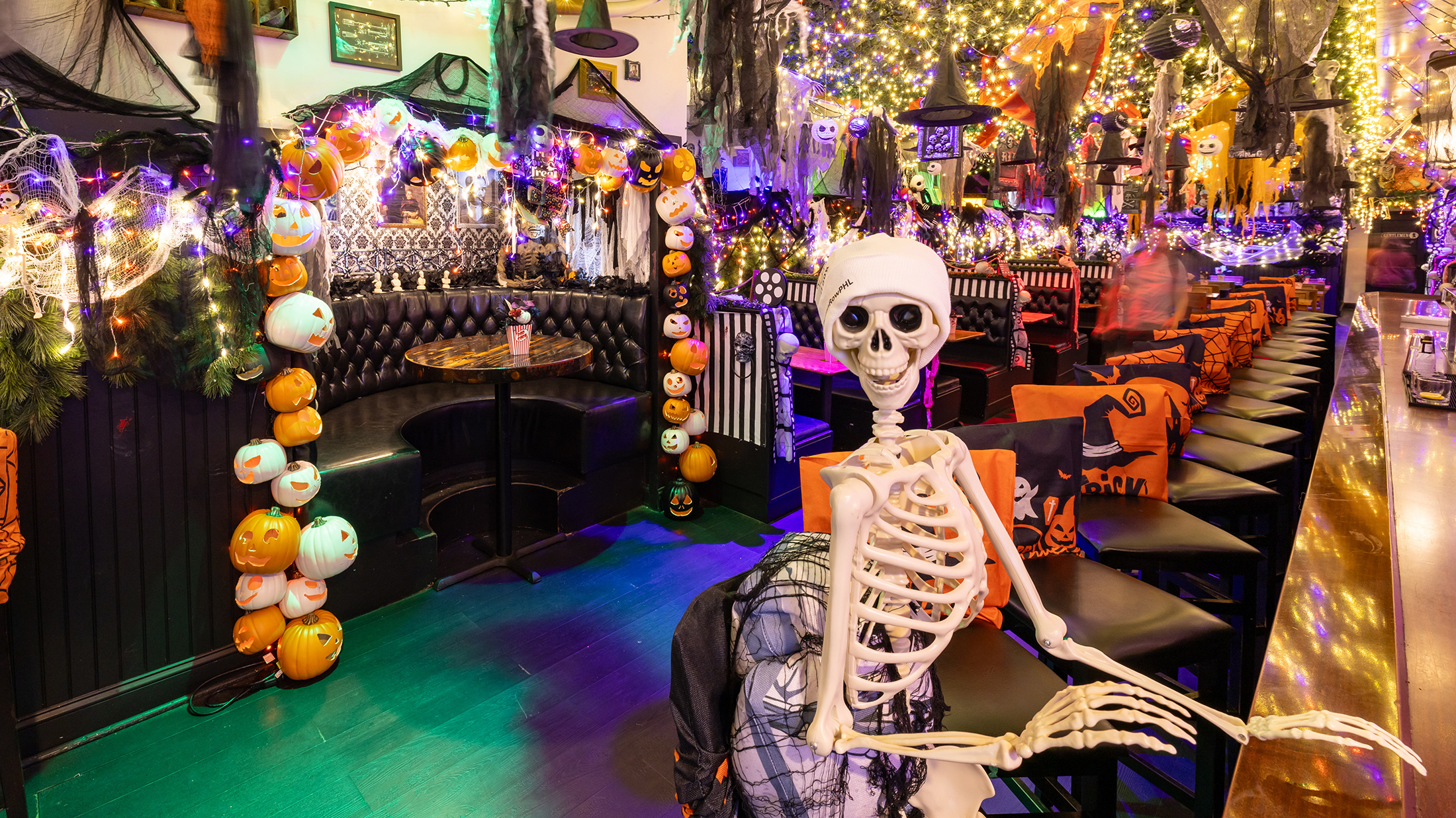 A plastic skeleton sits on a bar stool at a long wooden bar, surrounded by glowing pumpkin string lights, cobwebs, and colorful Halloween decorations hanging from the ceiling.