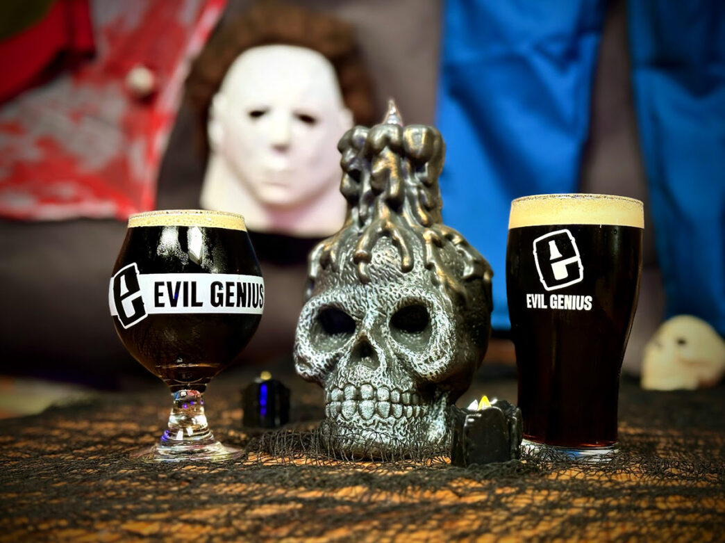 Two dark beers in branded Evil Genius glasses sit on a Halloween-themed table with a silver skull decoration in the center and a mask in the background.