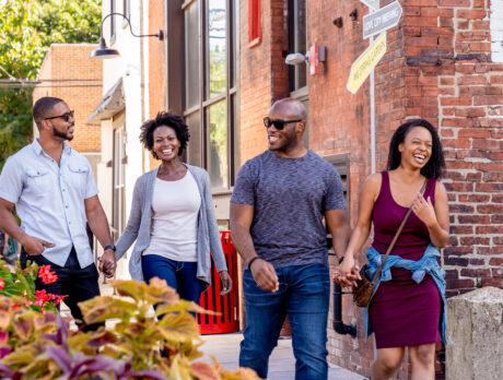 Four people laugh and walk hand-in-hand down a sidewalk lined with brick buildings on a sunny day.