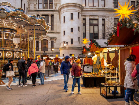 Visitors browse wooden vendor stalls decorated with garland and star-shaped lanterns and ride a carousel at Philadelphia's Christmas Village.