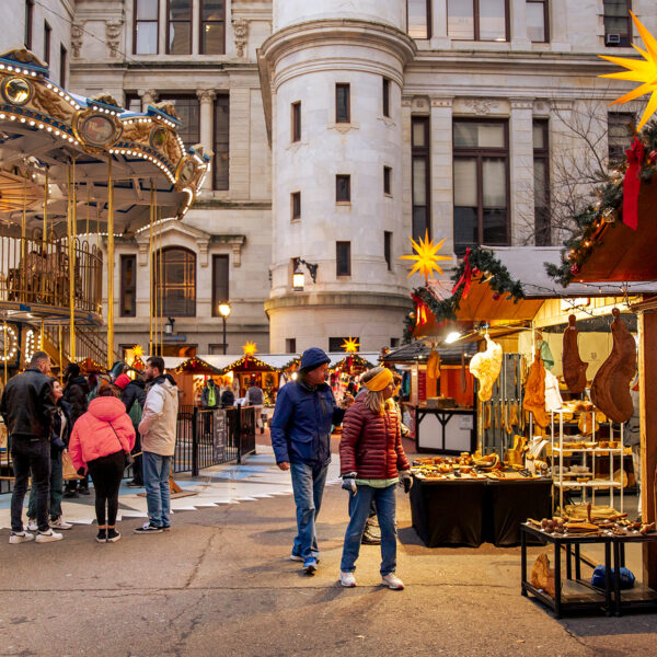 Visitors browse wooden vendor stalls decorated with garland and star-shaped lanterns and ride a carousel at Philadelphia's Christmas Village.