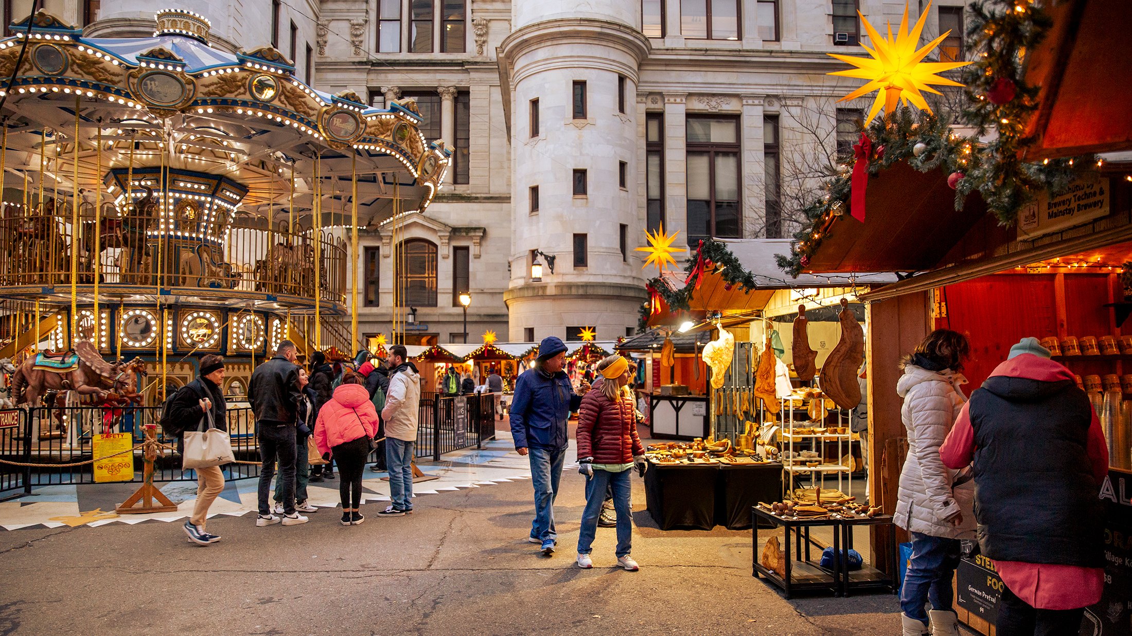 Visitors browse wooden vendor stalls decorated with garland and star-shaped lanterns and ride a carousel at Philadelphia's Christmas Village.