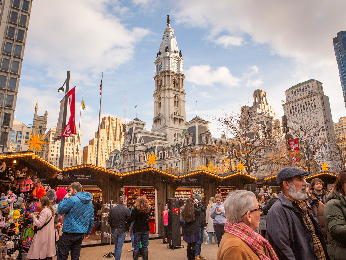 Crowds browse festive wooden stalls decorated with lights and star-shaped lanterns, with City Hall rising in the background.