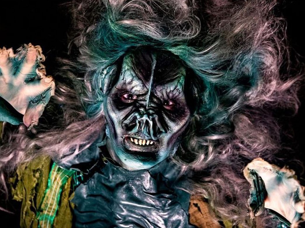 A performer in eerie makeup and a tattered costume snarls at the camera with webbed hands reaching outward at Eastern State Penitentiary.