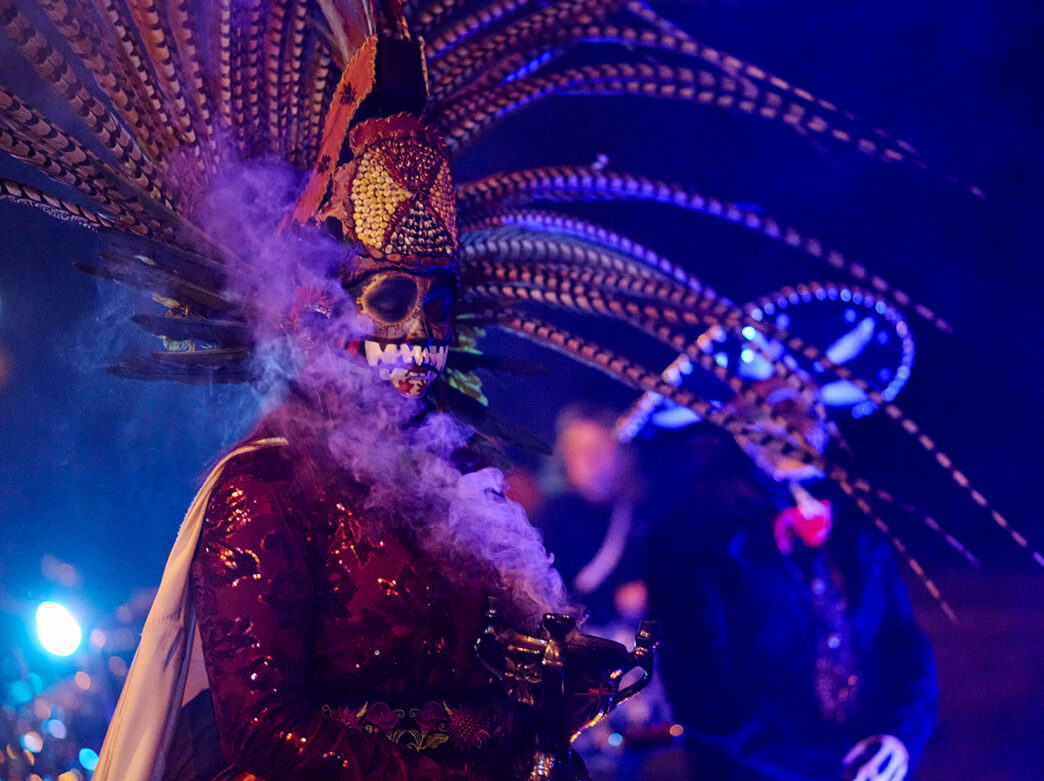 Un artista con un elaborado tocado de plumas y maquillaje de calavera participa en una celebración del Día de los Muertos en el Parque FDR.