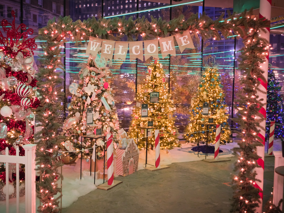 A festive indoor display of decorated Christmas trees shine with colorful lights, garland and a hanging "Welcome" sign.