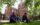 Three people sit in the grass outside Independence Hall, smiling and talking beneath the shade of leafy green trees.