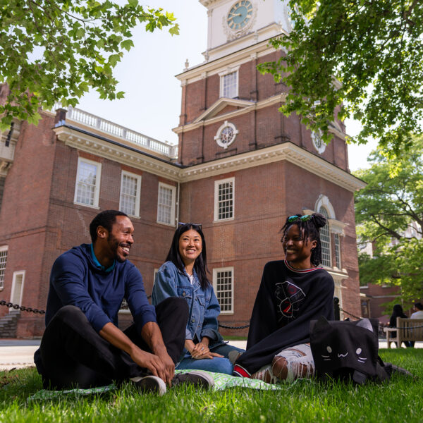 Trois personnes sont assises dans l'herbe à l'extérieur de Independence Hall, souriant et discutant à l'ombre d'arbres verts.