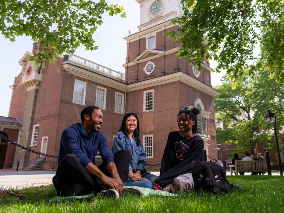 Three people sit in the grass outside Independence Hall, smiling and talking beneath the shade of leafy green trees.