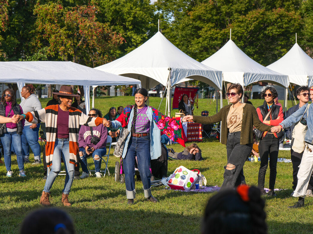 People join hands in a circle while dancing on the lawn at Indigenous Peoples Day, with white festival tents set up in the background.