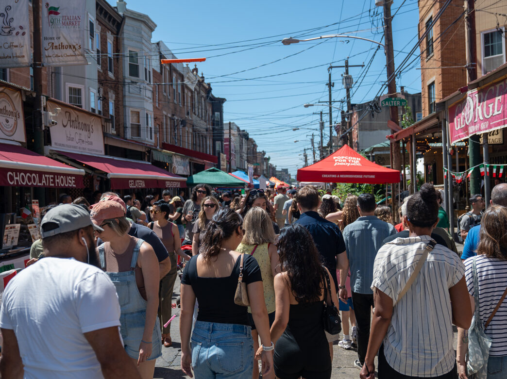 A crowd of people pack the South 9th Street Italian Market, browsing vendor stalls under colorful tents and signs during the Italian Market Festival.