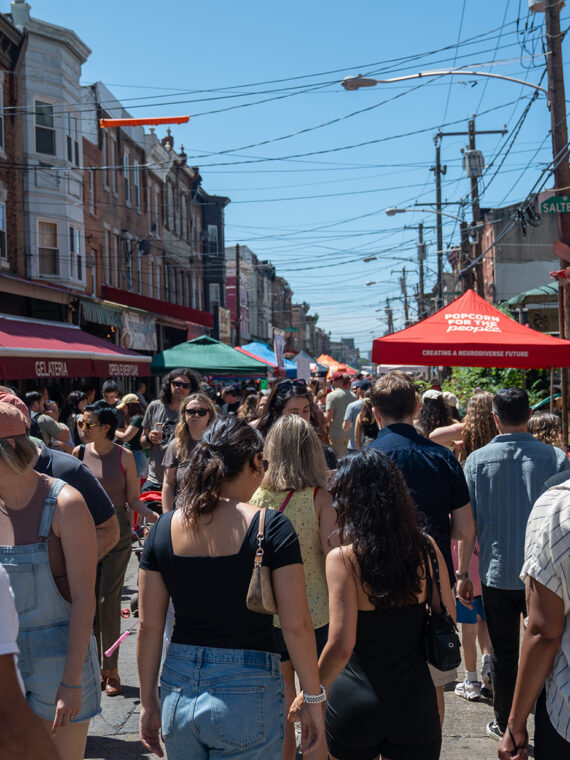 A crowd of people pack the South 9th Street Italian Market, browsing vendor stalls under colorful tents and signs during the Italian Market Festival.