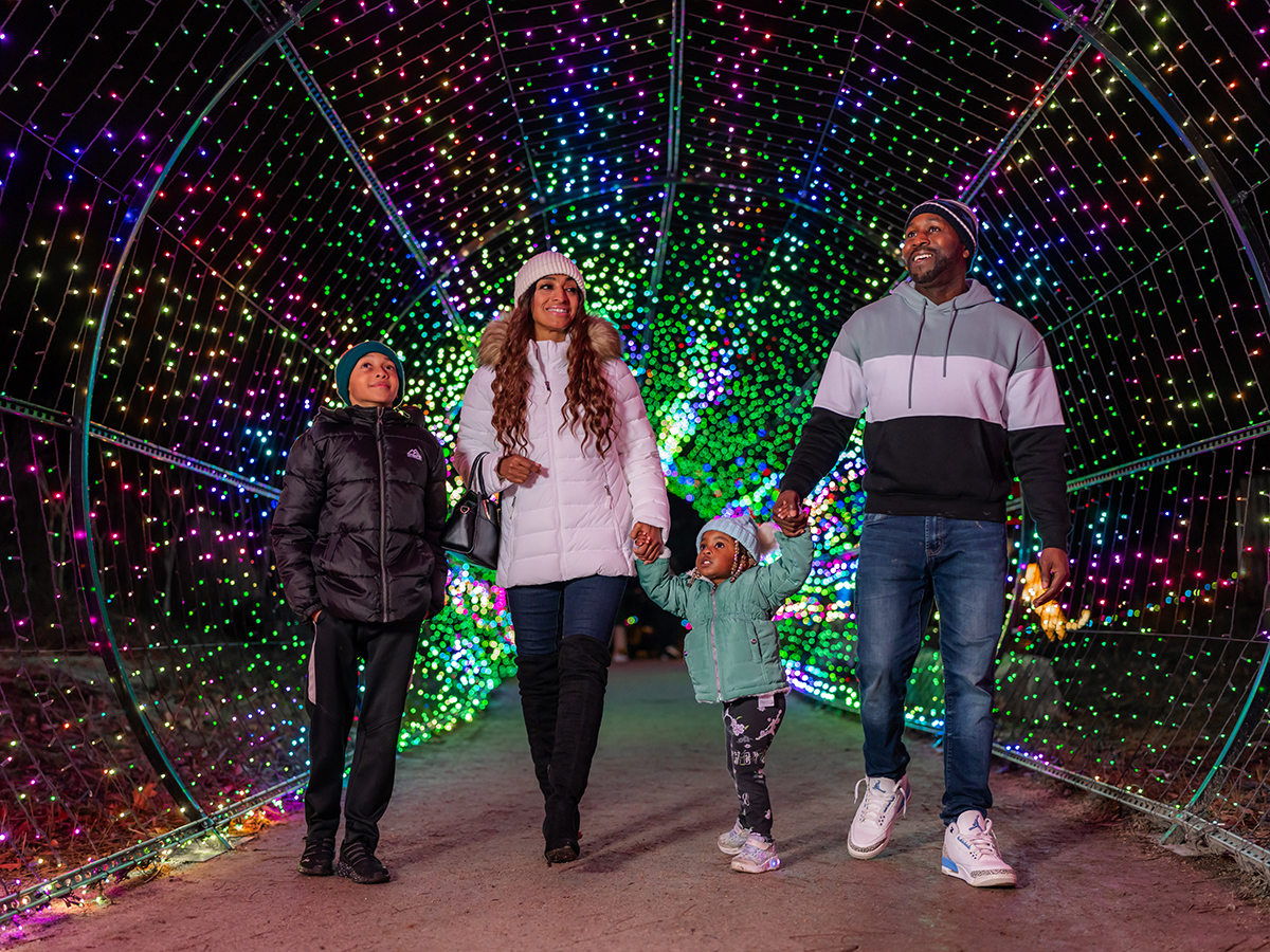 A family wearing winter jackets and beanies walks hand-in-hand through a tunnel of colorful holiday lights at night.