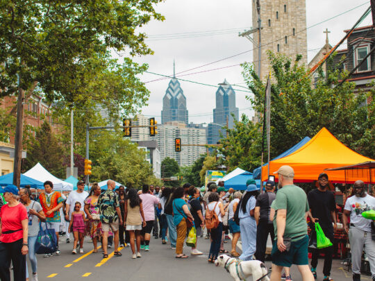 Pendant le festival Odunde, la foule défile dans une rue bordée de tentes colorées où s'affichent les stands des marchands, avec la silhouette de la ville en arrière-plan.