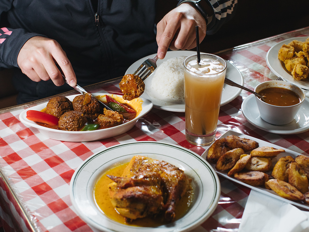 A person sits at a table with a red and white gingham tablecloth and enjoys Latin American dishes, including rice, fried plantains and stewed meat.