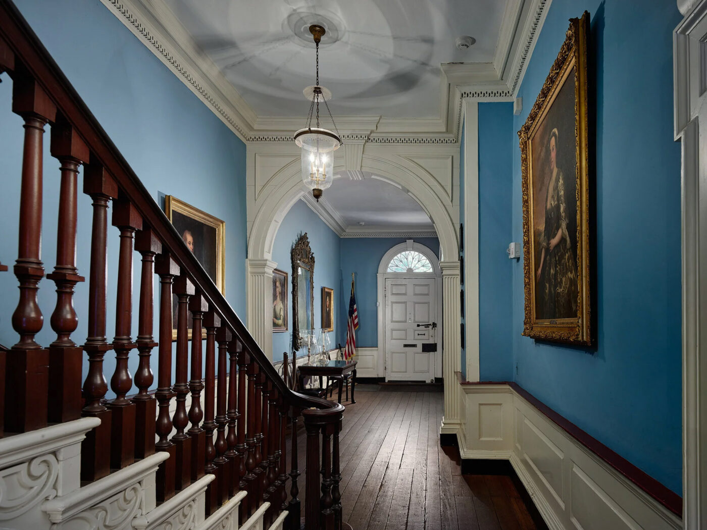 A photo of an elegant, blue hallway featuring a stately wooden staircase and gilded portraits inside the Powel House.