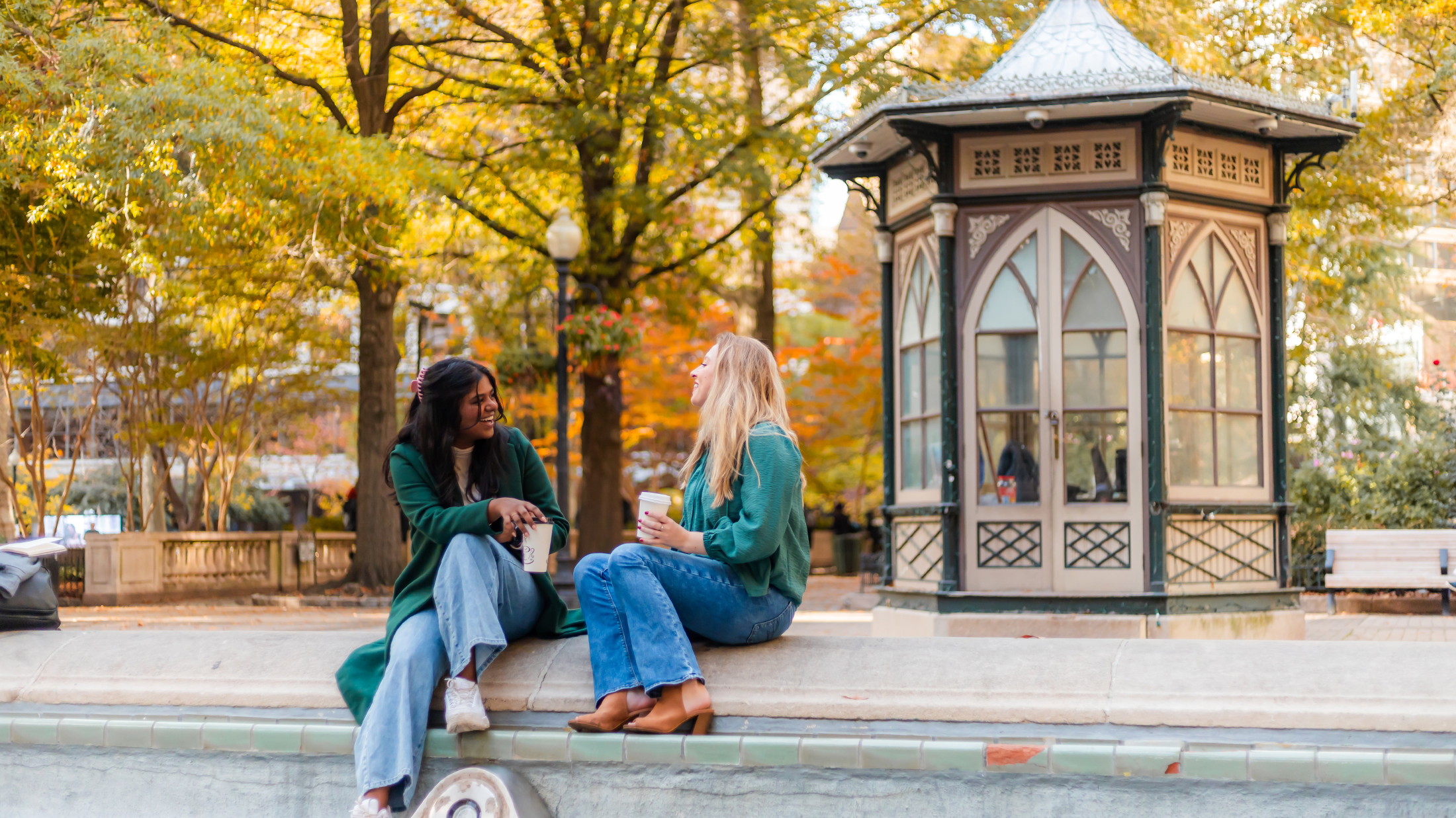 Two friends sit on the ledge of a drained fountain in Rittenhouse Square on a sunny autumn day, laughing and enjoying coffee beneath colorful fall foliage.
