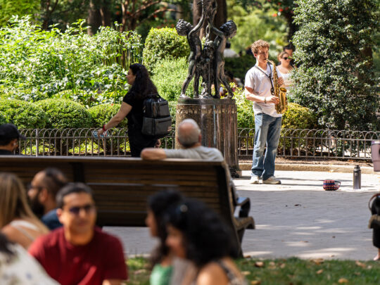 A saxophonist stands next to a sculpture in Rittenhouse Square and plays for a crowd of onlookers sitting on benches and the grass.