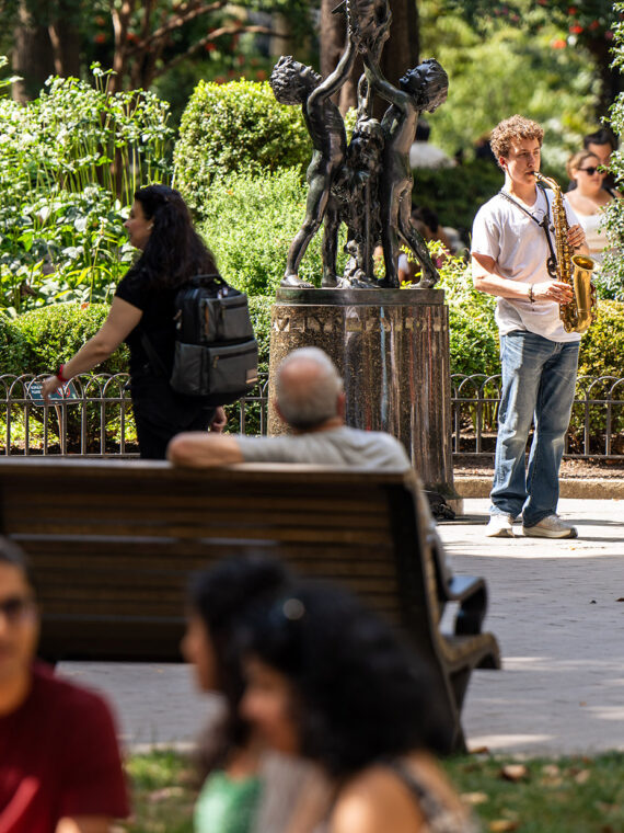 A saxophonist stands next to a sculpture in Rittenhouse Square and plays for a crowd of onlookers sitting on benches and the grass.