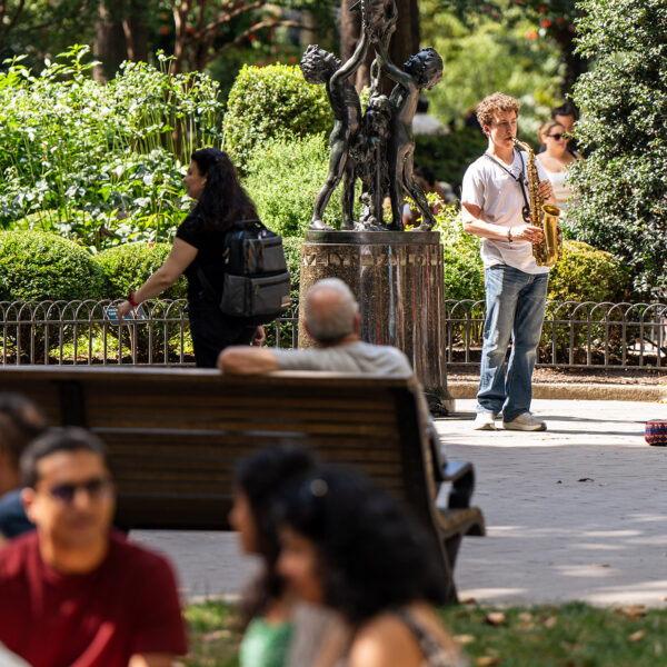 Een saxofonist staat naast een sculptuur in Rittenhouse Square en speelt voor een groep toeschouwers die op bankjes en op het gras zitten.