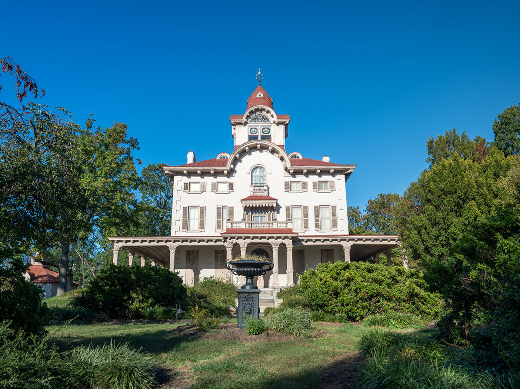 The exterior of Ryerss Museum and Liberty. A grand 19th-century mansion with a red roof and clock tour surrounded by lush greenery under a blue sky.