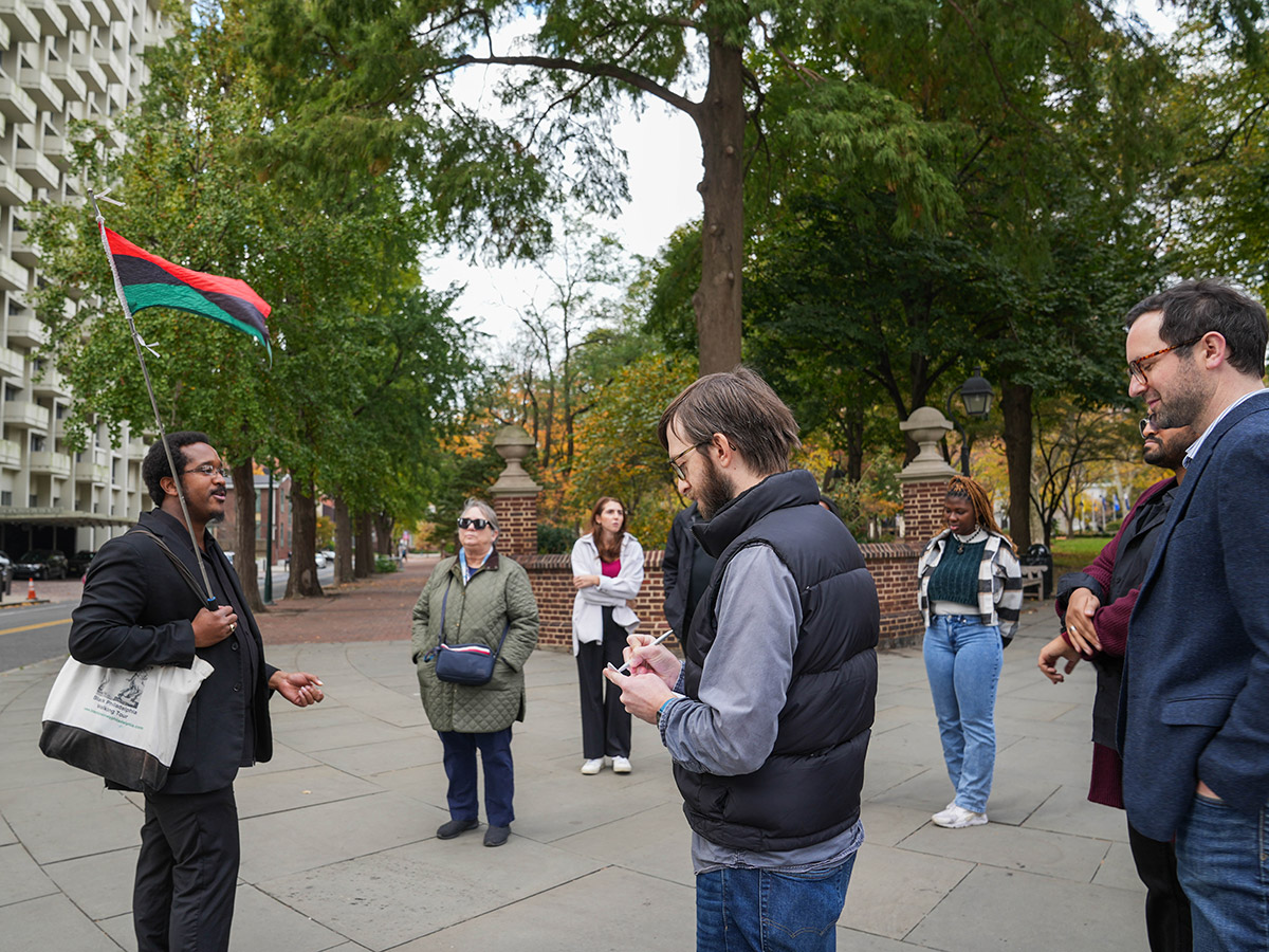 A tour guide for The Black Journey Walking Tour holds a red, black and green Pan-African flag and leads a group through Washington Square Park.