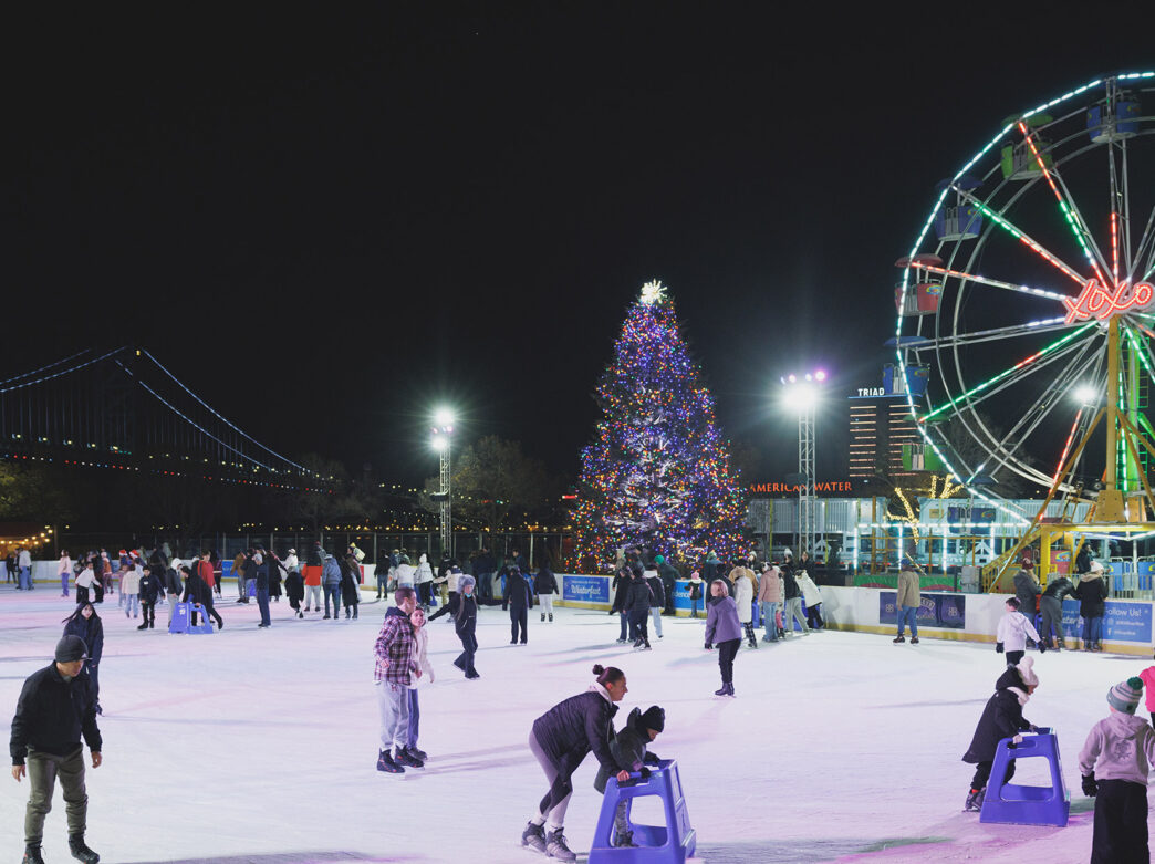 People ice skate on a white skating rink with colorful lights and a lit-up Ferris wheel in the background