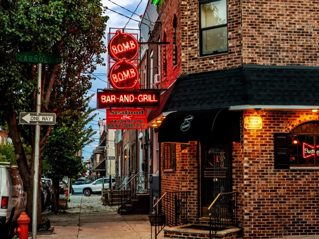 A neon red sign hangs from the side of a corner brick building.
