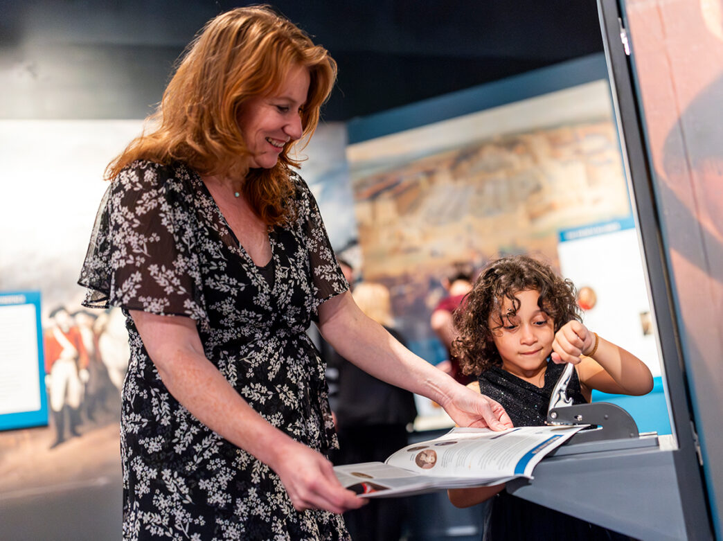 A woman and child engage in an interactive activity at the Museum of the American revolution, using a hand press to stamp a booklet.