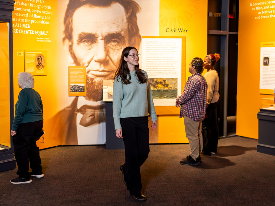 Visitors explore a new exhibit at the Museum of the American Revolution featuring artifacts, text panels and a large image of Abraham Lincoln in the gallery.