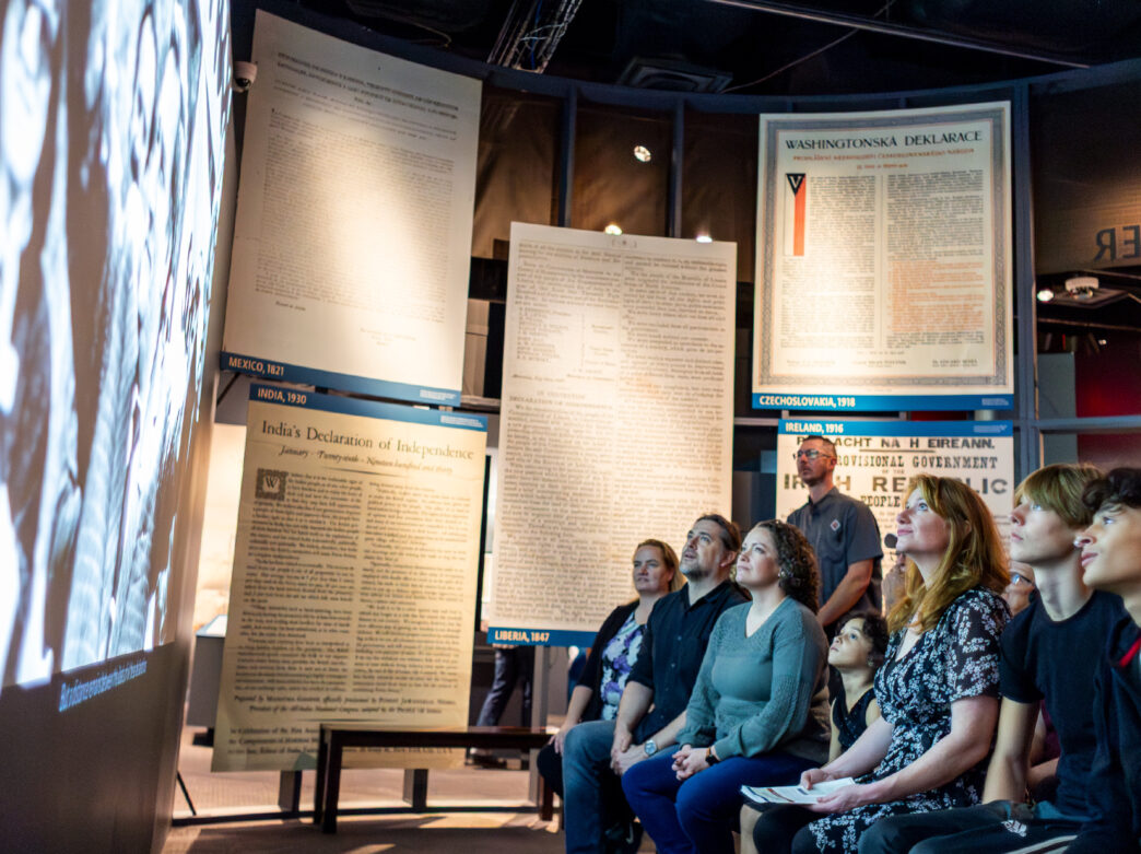 A group of visitors watch a film inside a museum exhibit, surrounded by reproductions of global declarations of independence.
