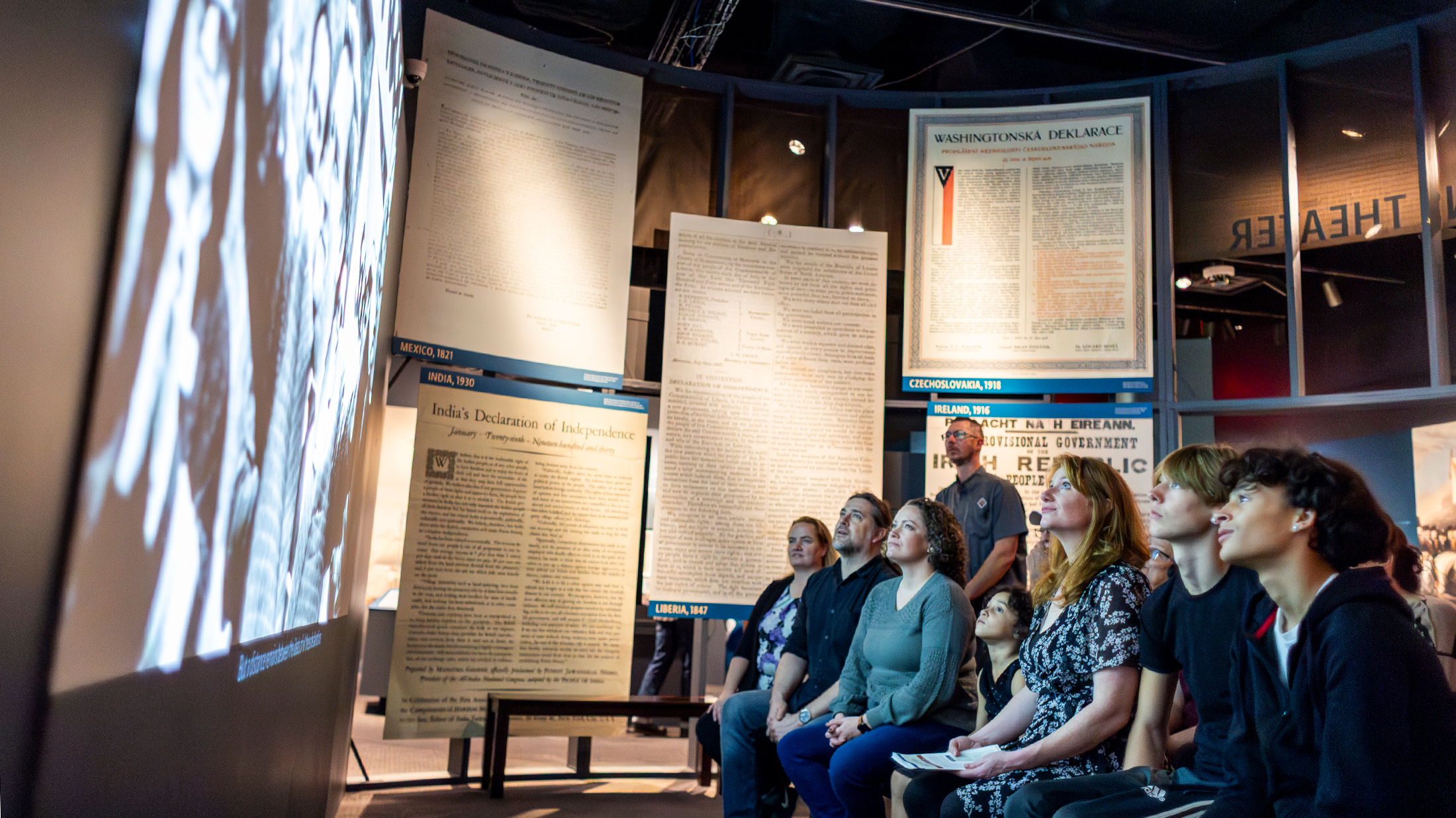 Un grupo de visitantes ve una película dentro de una exposición del museo, rodeados de reproducciones de declaraciones de independencia de todo el mundo.