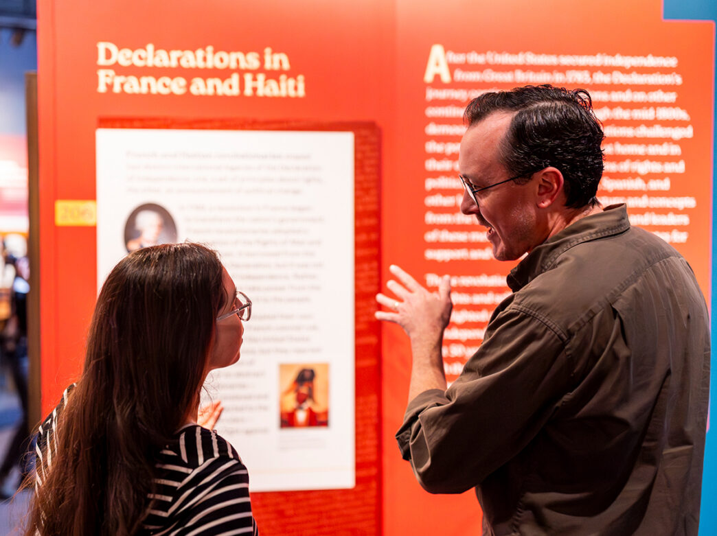 Dos visitantes se miran y conversan frente a un expositor rojo con el texto "Declaraciones en Francia y Haití".