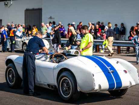 A crowd gathers outdoors around a white vintage racing car with blue stripes as museum staff inspect it during a demo event at the Simeone Foundation Automotive Museum.