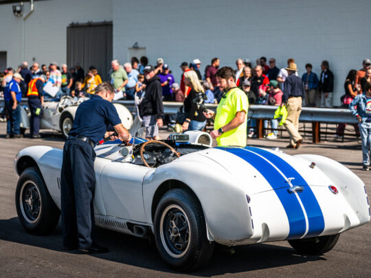 A crowd gathers outdoors around a white vintage racing car with blue stripes as museum staff inspect it during a demo event at the Simeone Foundation Automotive Museum.