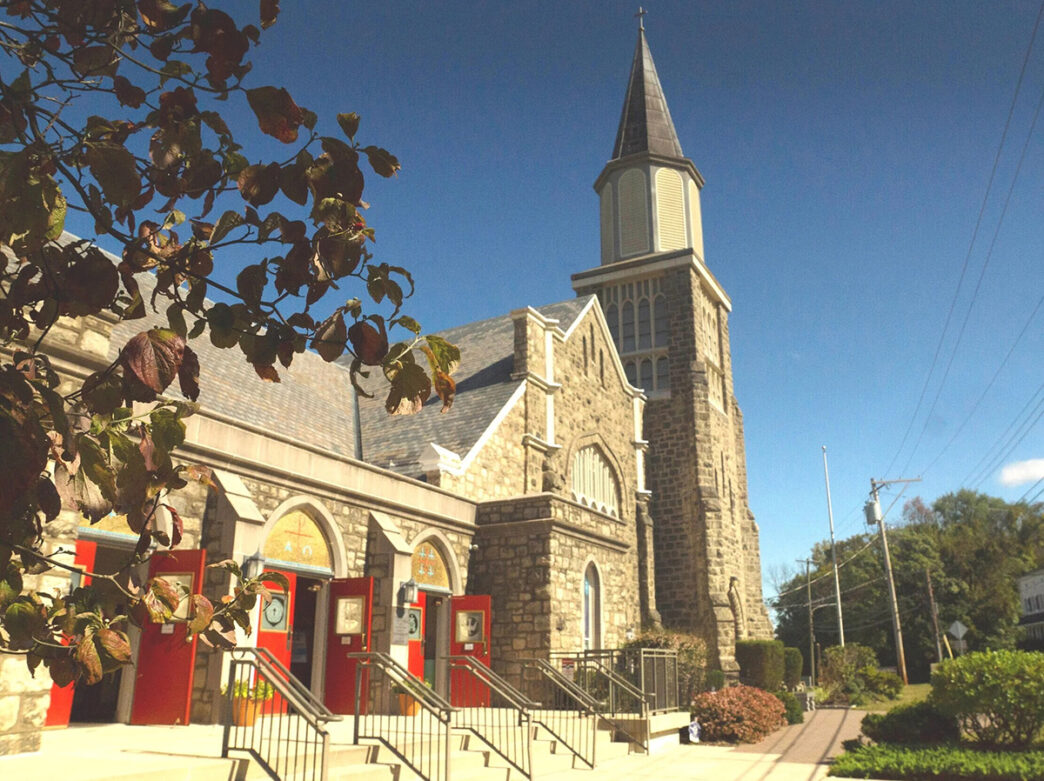 Exterior of a church in Lafayette Hill, with a stone facade, red doors and tall steeple against a clear blue sky.