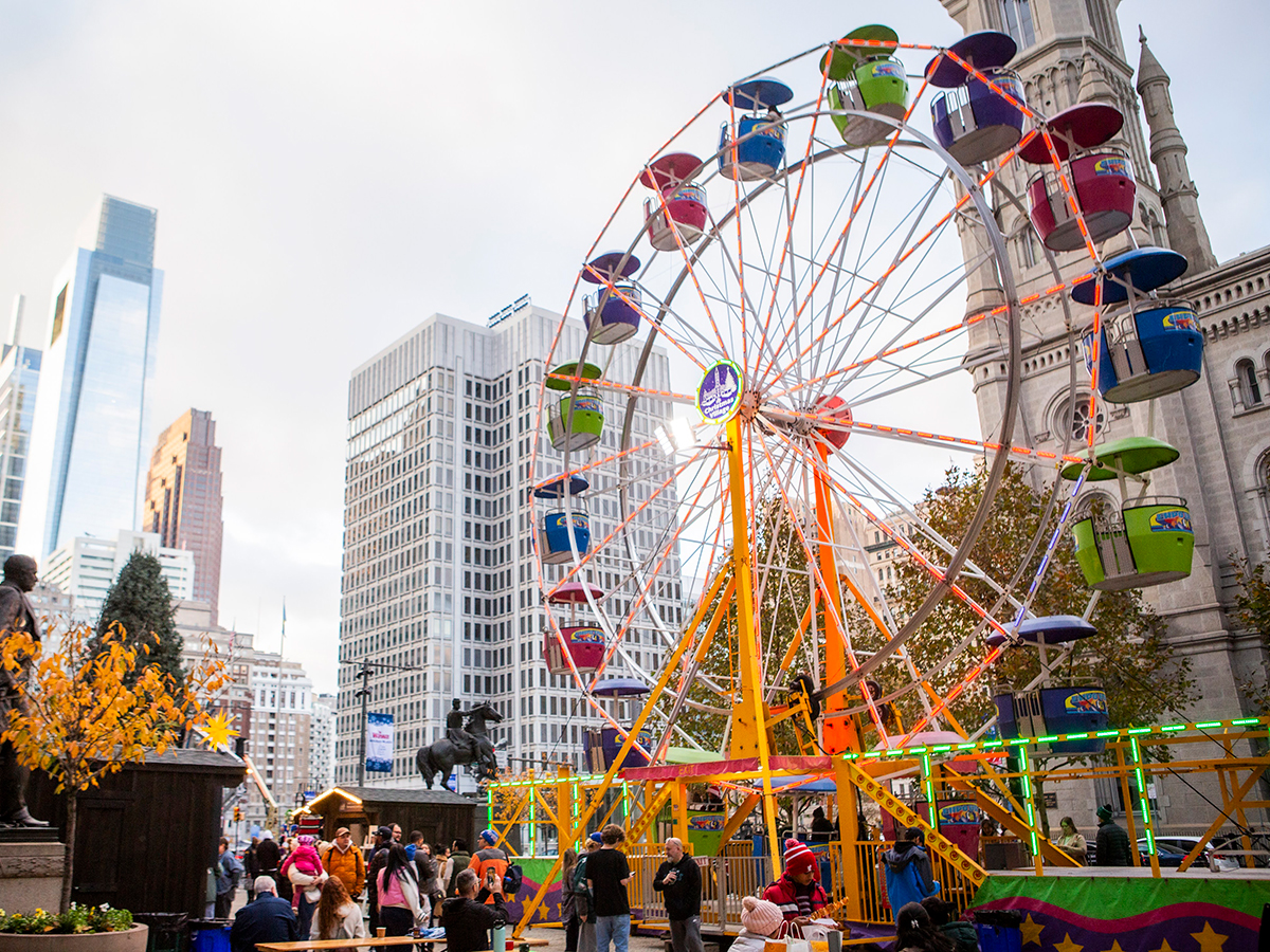 Visitors ride a colorful Ferris wheel surrounded by tall buildings and trees near Philadelphia's City Hall.