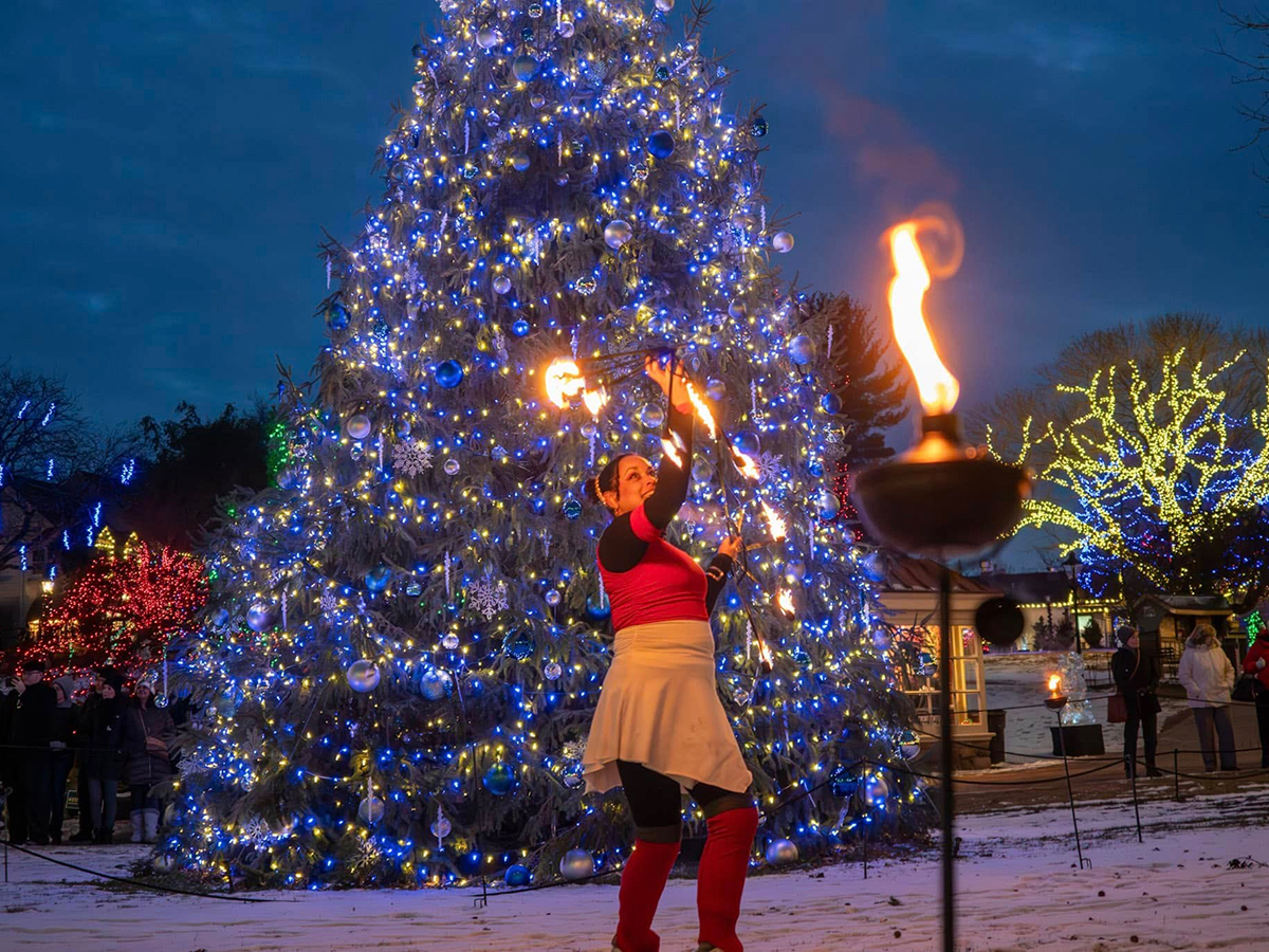 A performer spins flaming batons in front of a towering Christmas tree lit with blue and gold lights at Peddler's Village.