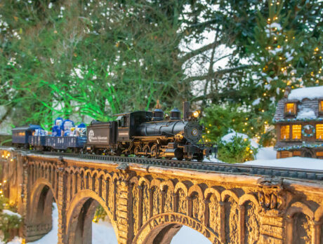 A miniature holiday train travels across a bridge surrounded by snow, twinkling lights and evergreen trees at Morris Arboretum's Garden Railway display.
