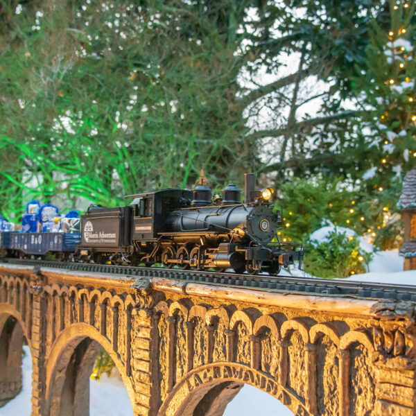 A miniature holiday train travels across a bridge surrounded by snow, twinkling lights and evergreen trees at Morris Arboretum's Garden Railway display.