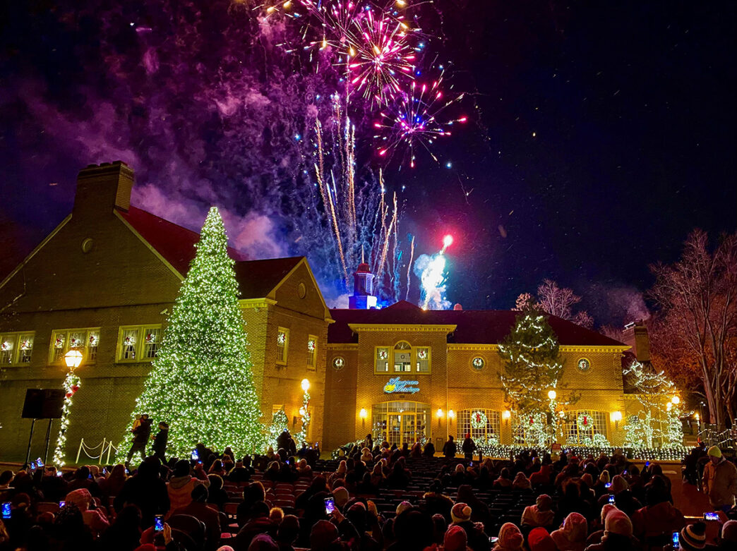 Crowds gather outside a festively lit brick building to watch fireworks explode in the sky above a massive Christmas tree.